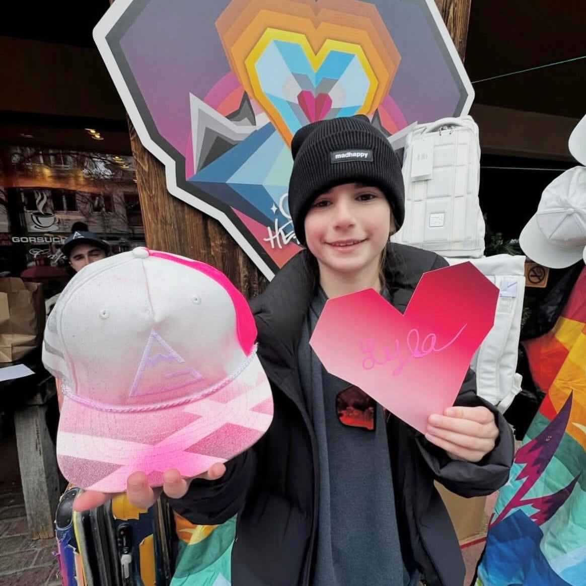 Child holding a pink cap and heart-shaped sign in front of colorful artwork.