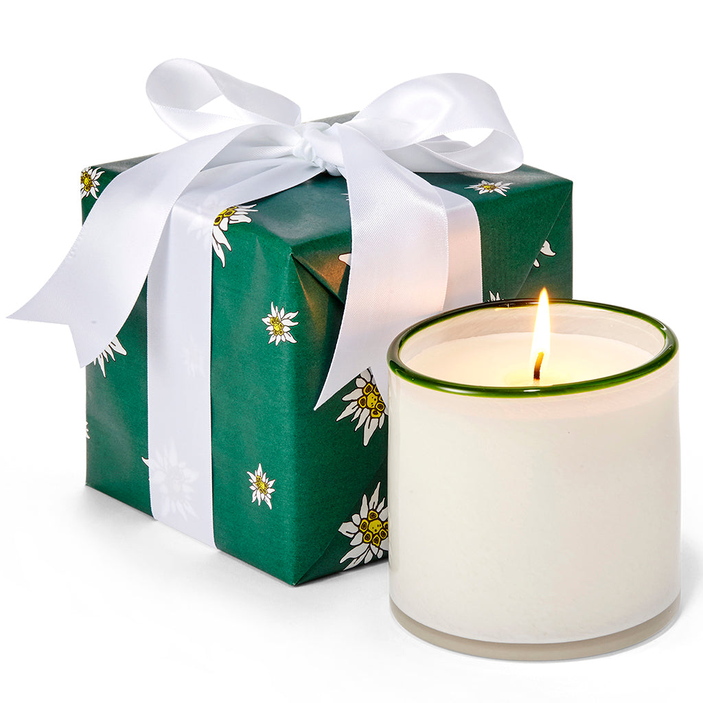 Exclusive for Gorsuch, the Alpine Fir candle, a white candle with green trim at the top, sits next to a gift box wrapped in green and white edelweiss paper with a white ribbon.