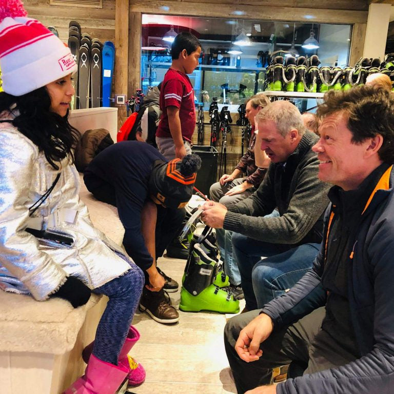 Young children with adults in a ski shop with skis and equipment in the background.