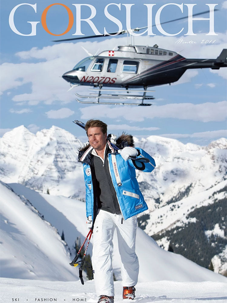 2011 Gorsuch catalog cover with man in turquoise ski jacket carrying skis and poles with a helicopter above him and mountain range in the background.