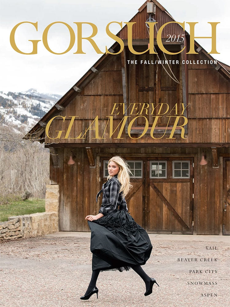 2015 Gorsuch catalog cover with woman in a plaid blouse and black taffeta skirt and heels prancing in front of a wood barn garage.