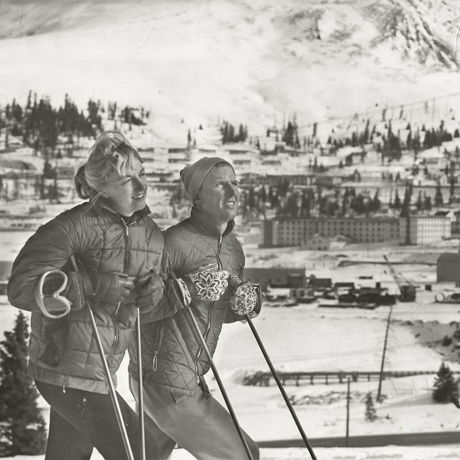 David and Renie Gorsuch standing on skis at the top of a mountain, holding ski poles and surrounded by a scenic alpine backdrop.
