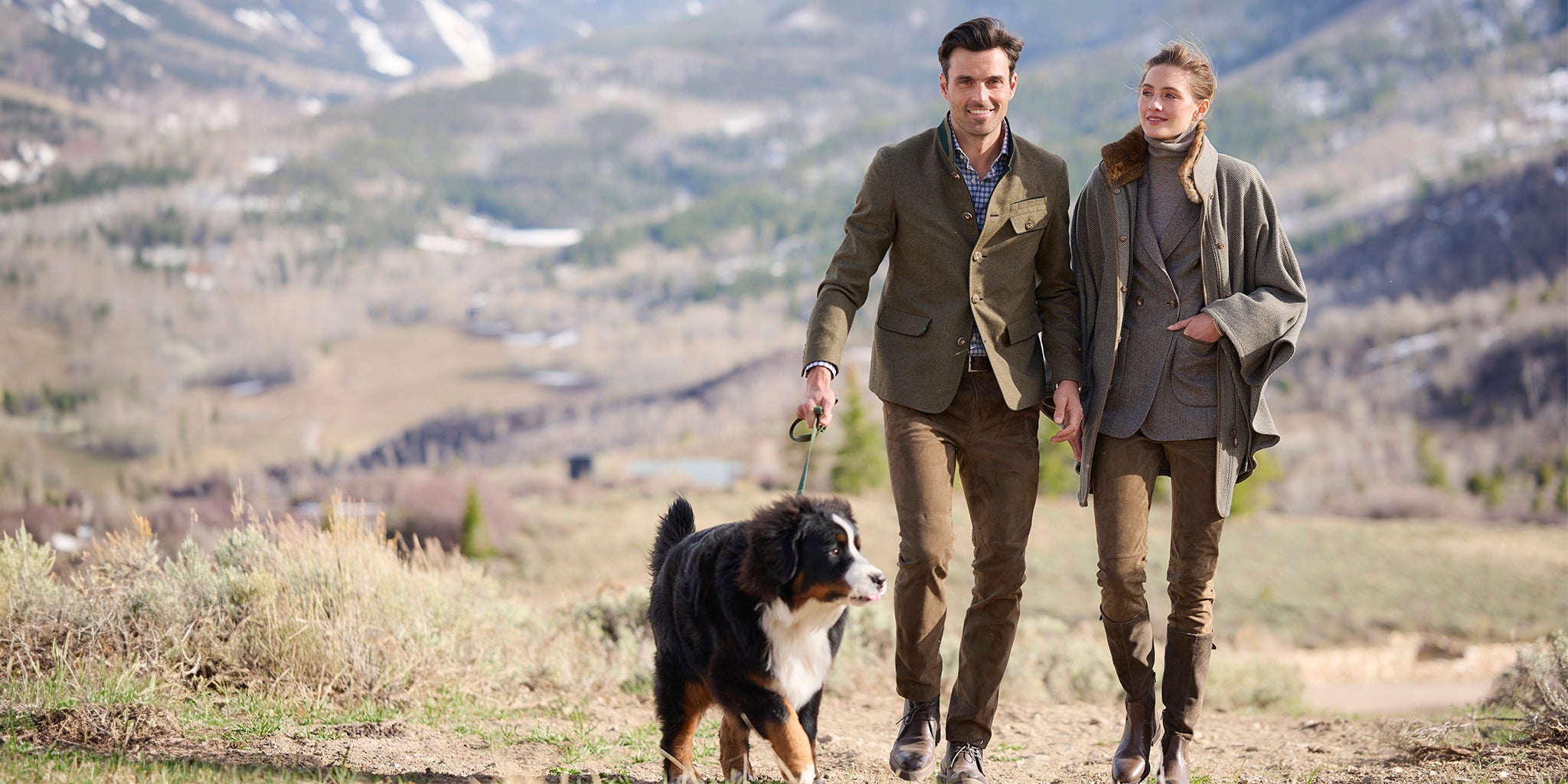 Couple walking their Bernese Mountain Dog outdoors, with the woman wearing the grey Kolbenturm Wool Cape and the man in the green Jakob Wool Jacket, set against a mountainous landscape.