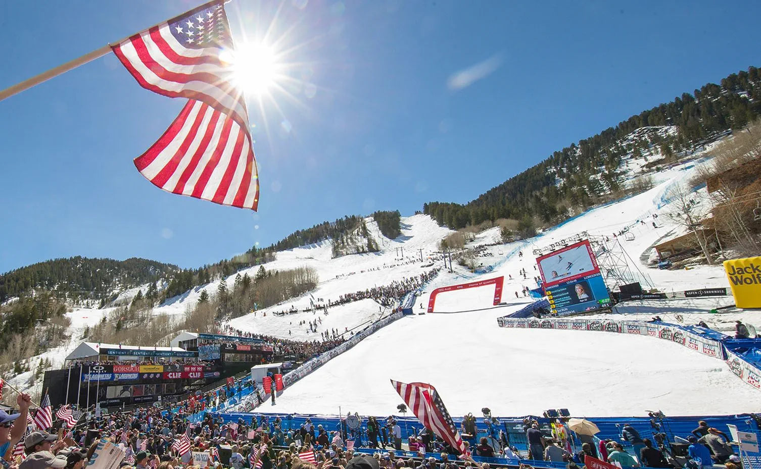 View from the bottom of a ski race course, showcasing a large crowd gathered around, with the American flag waving prominently.