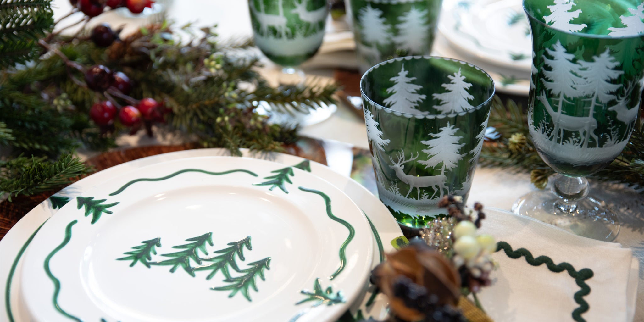 Festive holiday table setting with white plates featuring green trees and green glasses etched with deer and trees.