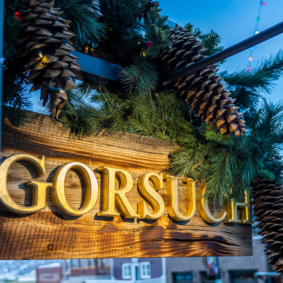 Decorative sign with 'GORSUCH' in gold letters, surrounded by pinecones and greenery against a blue sky.