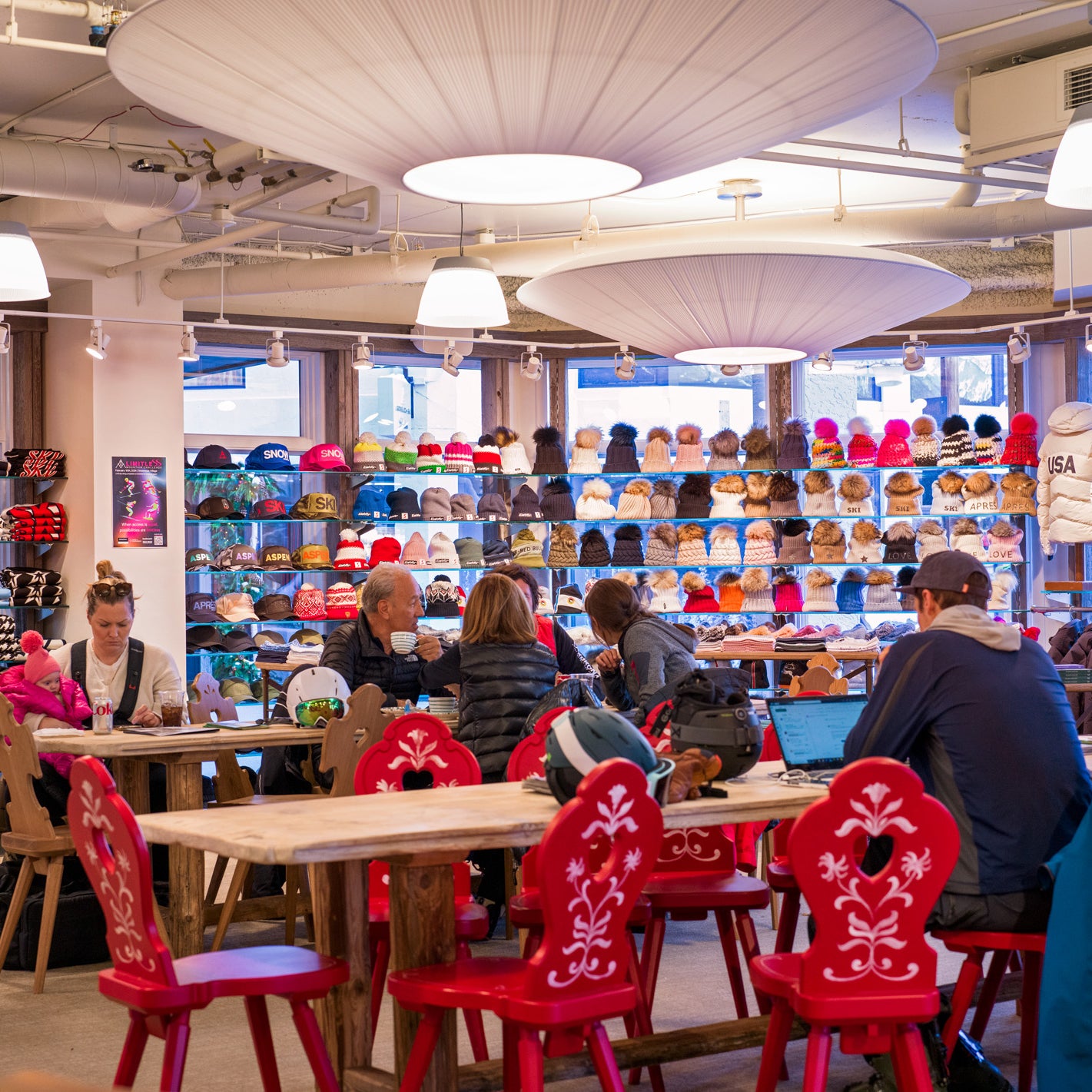 People sitting at tables with red chairs in the Snowmass Ski Cafe with shelves displaying various items.