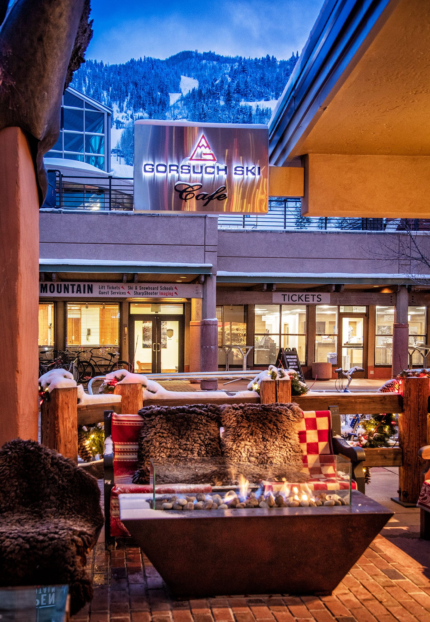 Outdoor seating area with a fire pit in front of a building with 'Gorsuch Ski Cafe' branding, Aspen mountain view in the background.