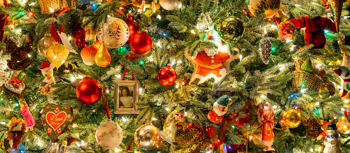 A close-up of a decorated Christmas tree with many ornaments, including red and gold baubles, string lights, a small framed picture, a Santa figure, pinecones, and other festive ornaments filling the frame. "The Holiday Gift Guide".