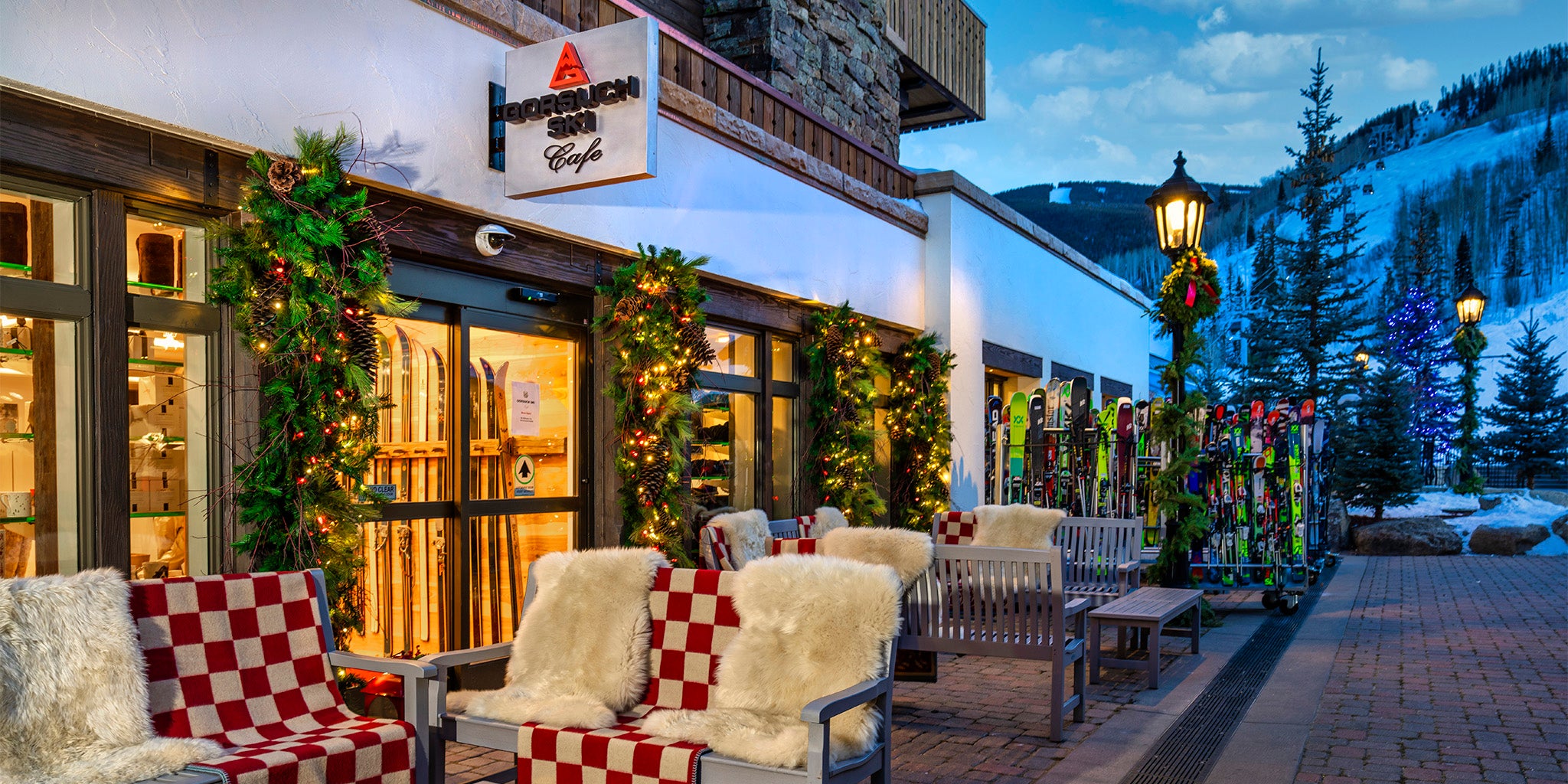 Outdoor seating area in front of the Gorsuch Ski Cafe in Vail with Christmas decorations, including garlands and lights, with a snowy mountain landscape in the background.