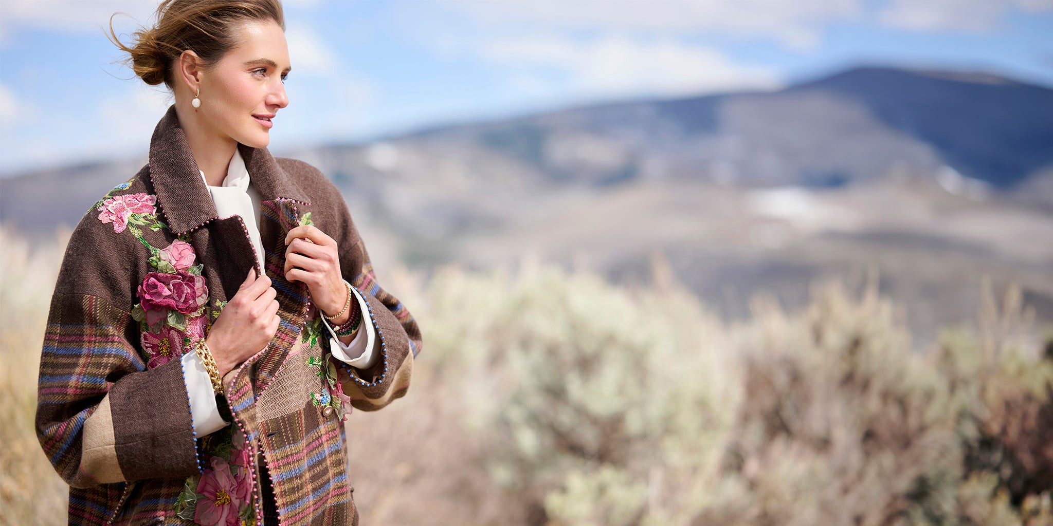 Woman wearing the Eliana Embroidered Plaid Coat by Péro, featuring a plaid and floral pattern, standing in front of a mountainous landscape.
