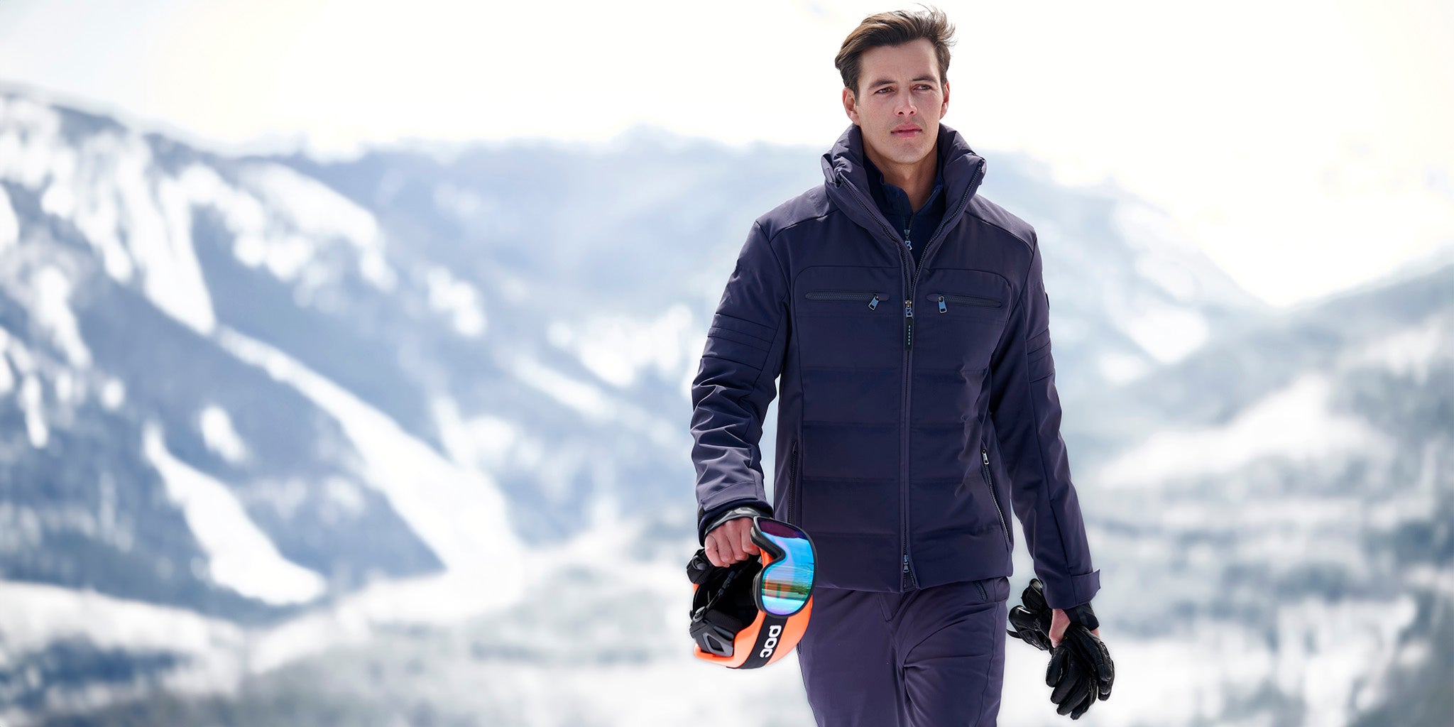 Man wearing the Minho Softshell Ski Jacket, holding ski gloves and a helmet against a snowy mountain background.