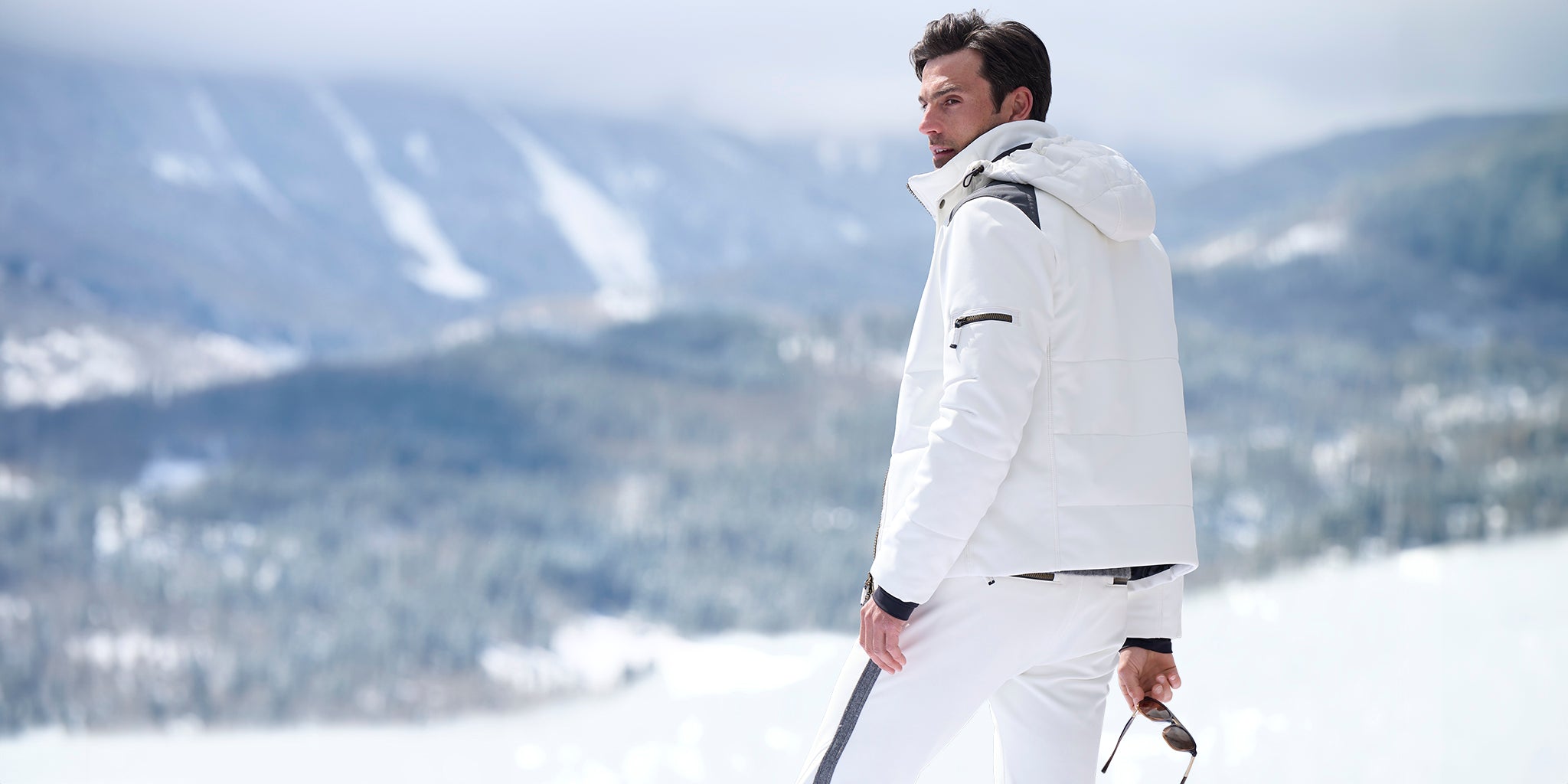 Man in white ski suit standing on a snowy slope with mountains in the background