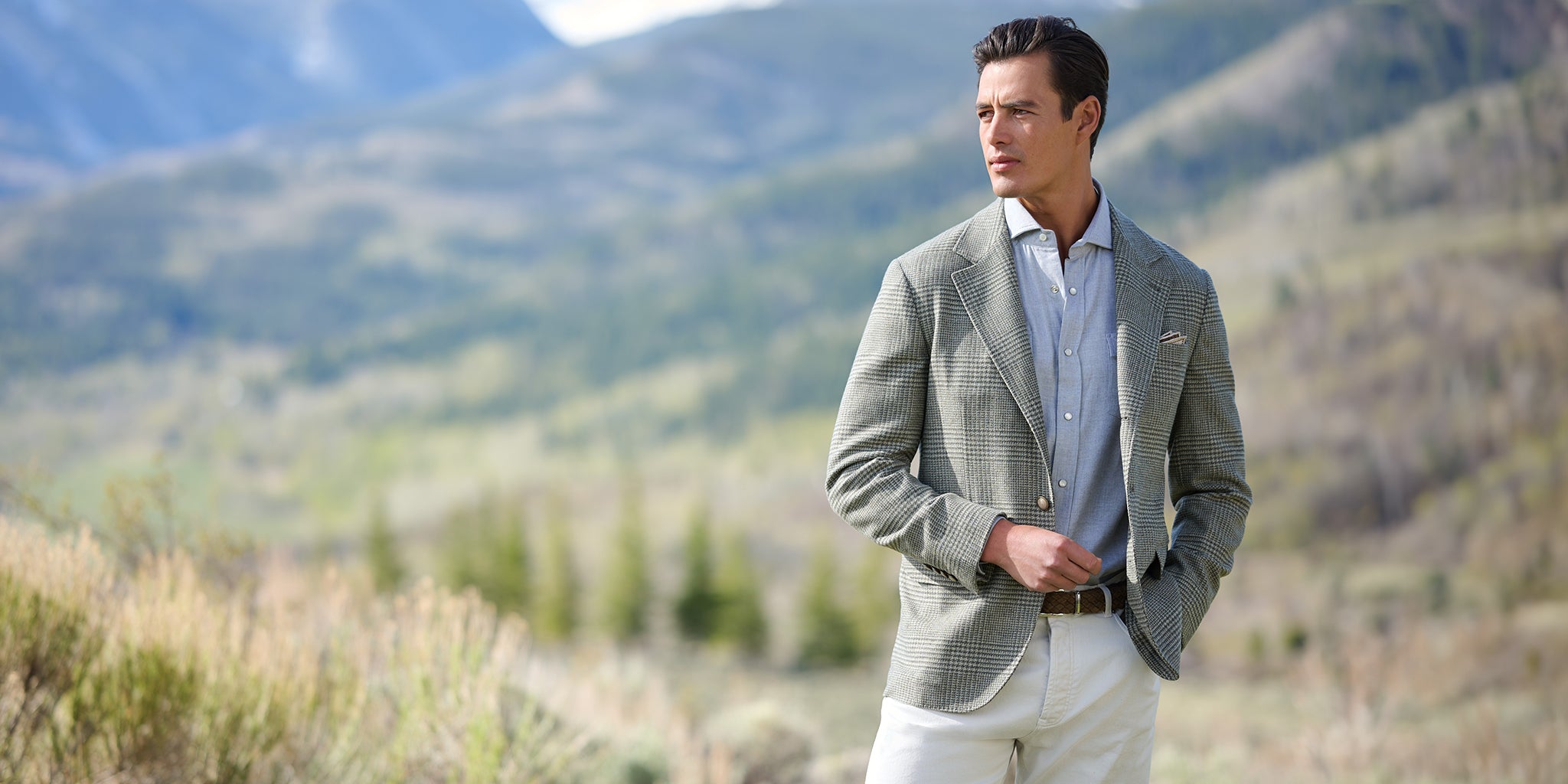 A man wearing the Prince of Wales Check Wool Blazer by Brunello Cucinelli, light grey Western Flannel Shirt and cream trousers stands outdoors against a blurred mountain landscape.