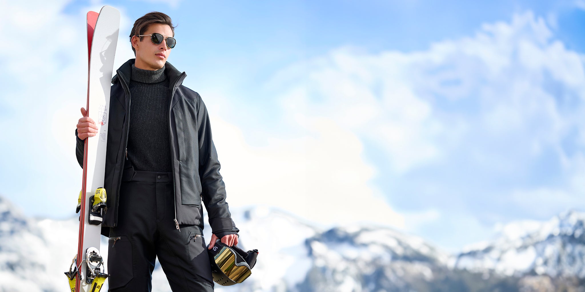 Man wearing the Trace Ski Jacket by Sease in carbon, holding skis in one hand and a helmet with goggles in the other, with snowy mountains in the background.