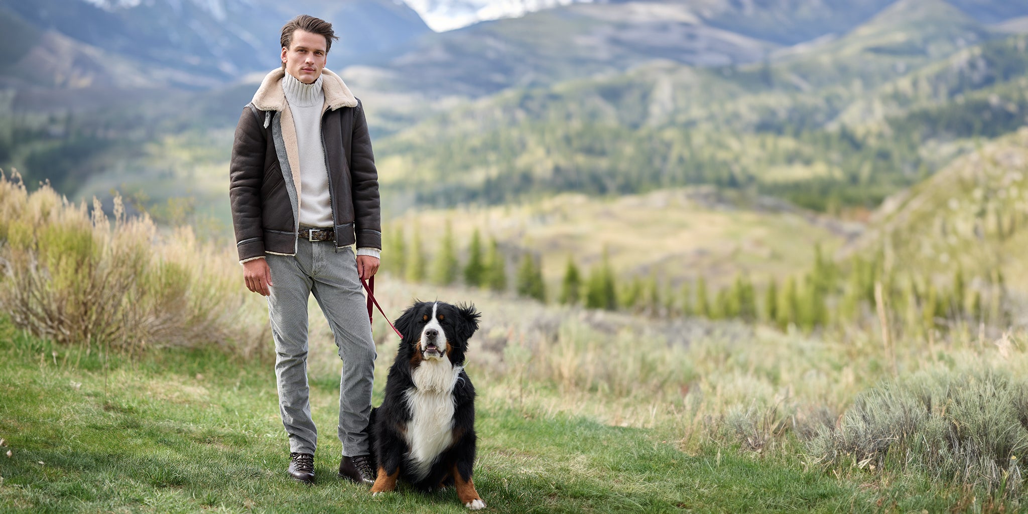 A man wearing a dark brown leather aviator jacket with a beige shearling collar, gray pants, and a white turtleneck stands outdoors in a mountain landscape next to a Bernese mountain dog.