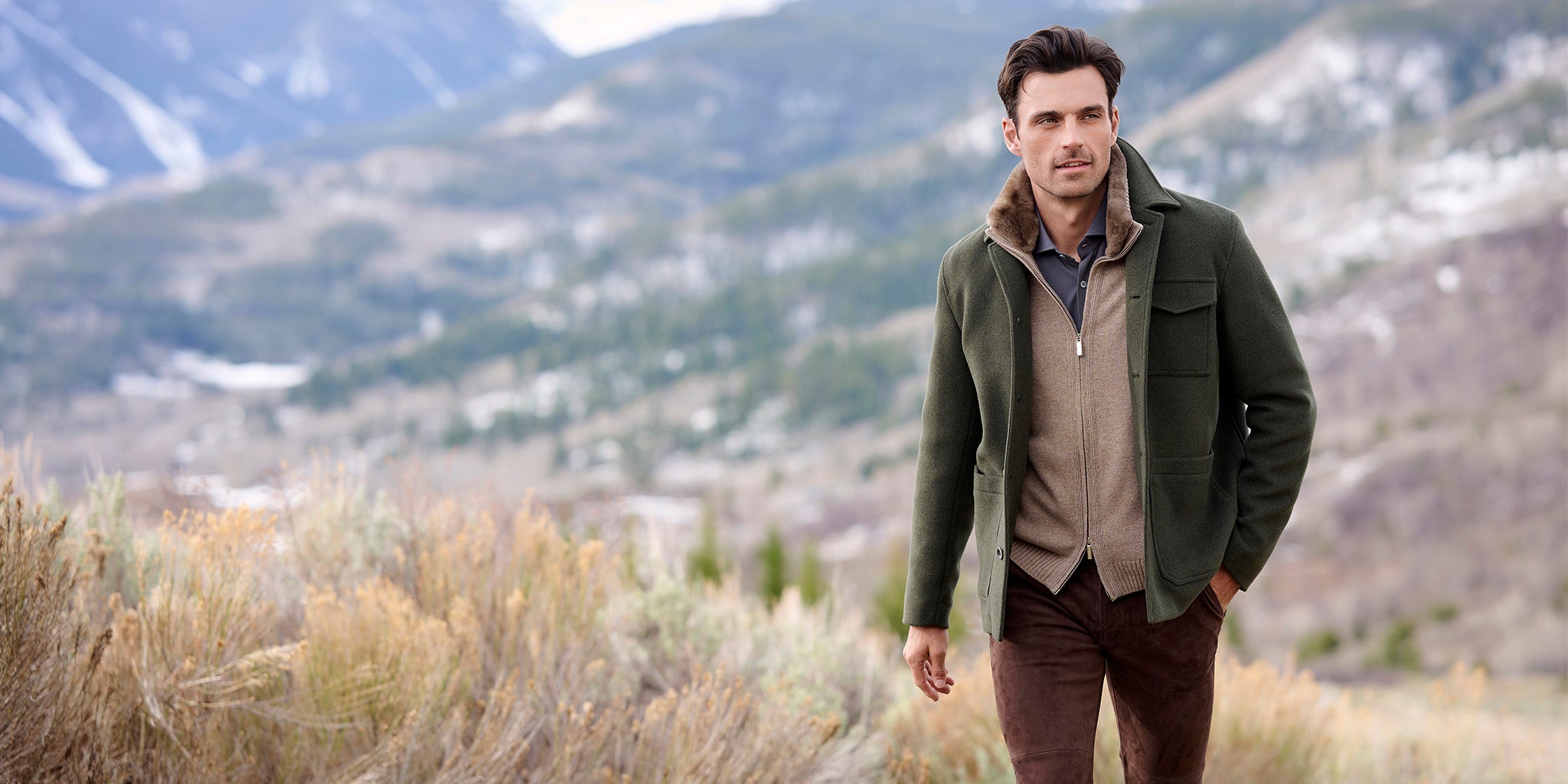 Man wearing a green Herno Brady Barn Coat layered over a brown zip cardigan, standing in front of a mountainous landscape.