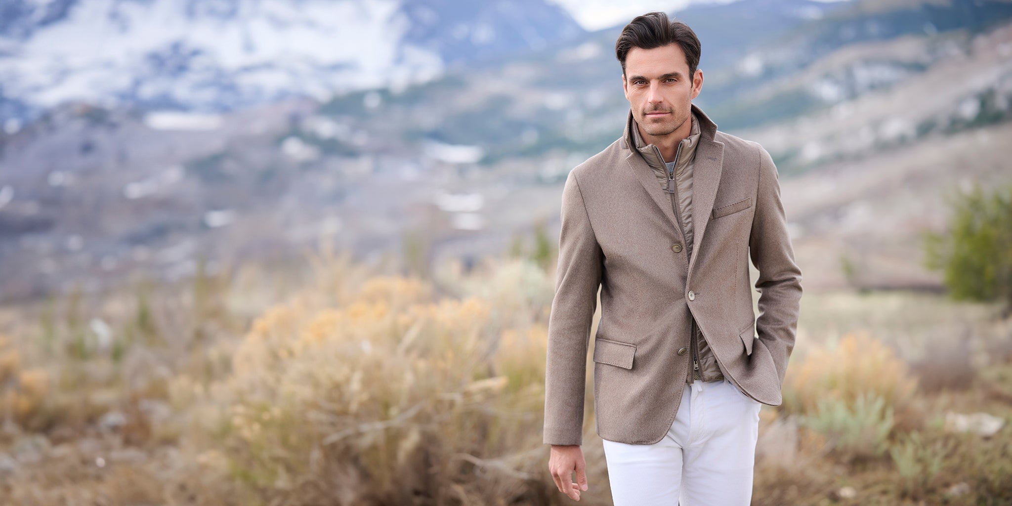 Man in a beige blazer and white pants standing in a scenic outdoor setting with mountains.
