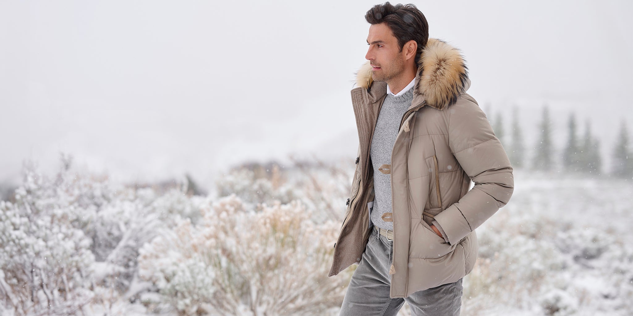 Man wearing a beige Arctic Down parka with a fur-trimmed hood, standing in a snowy landscape.