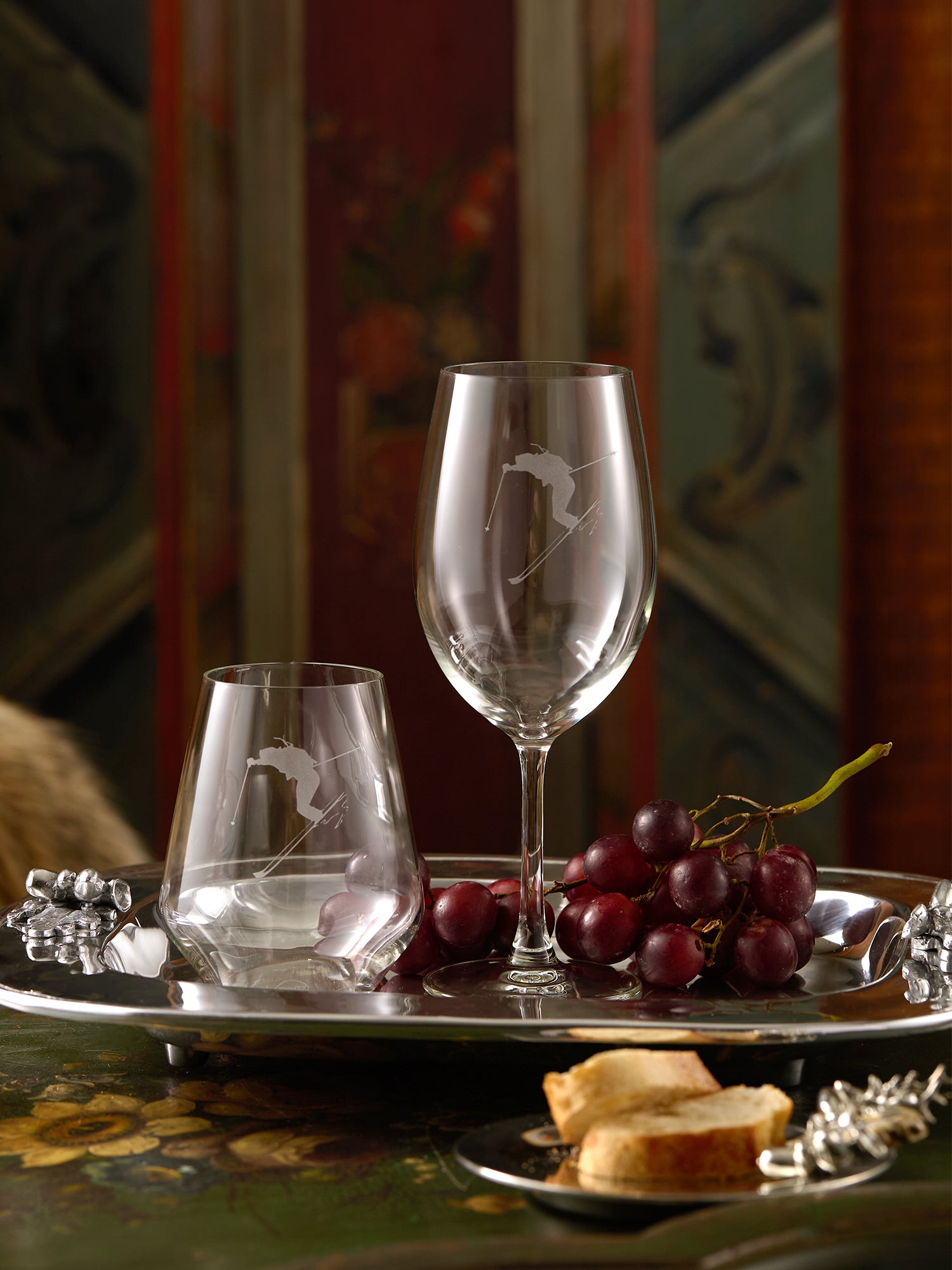 Two wine glasses with engraved skier designs on a tray with grapes and bread.