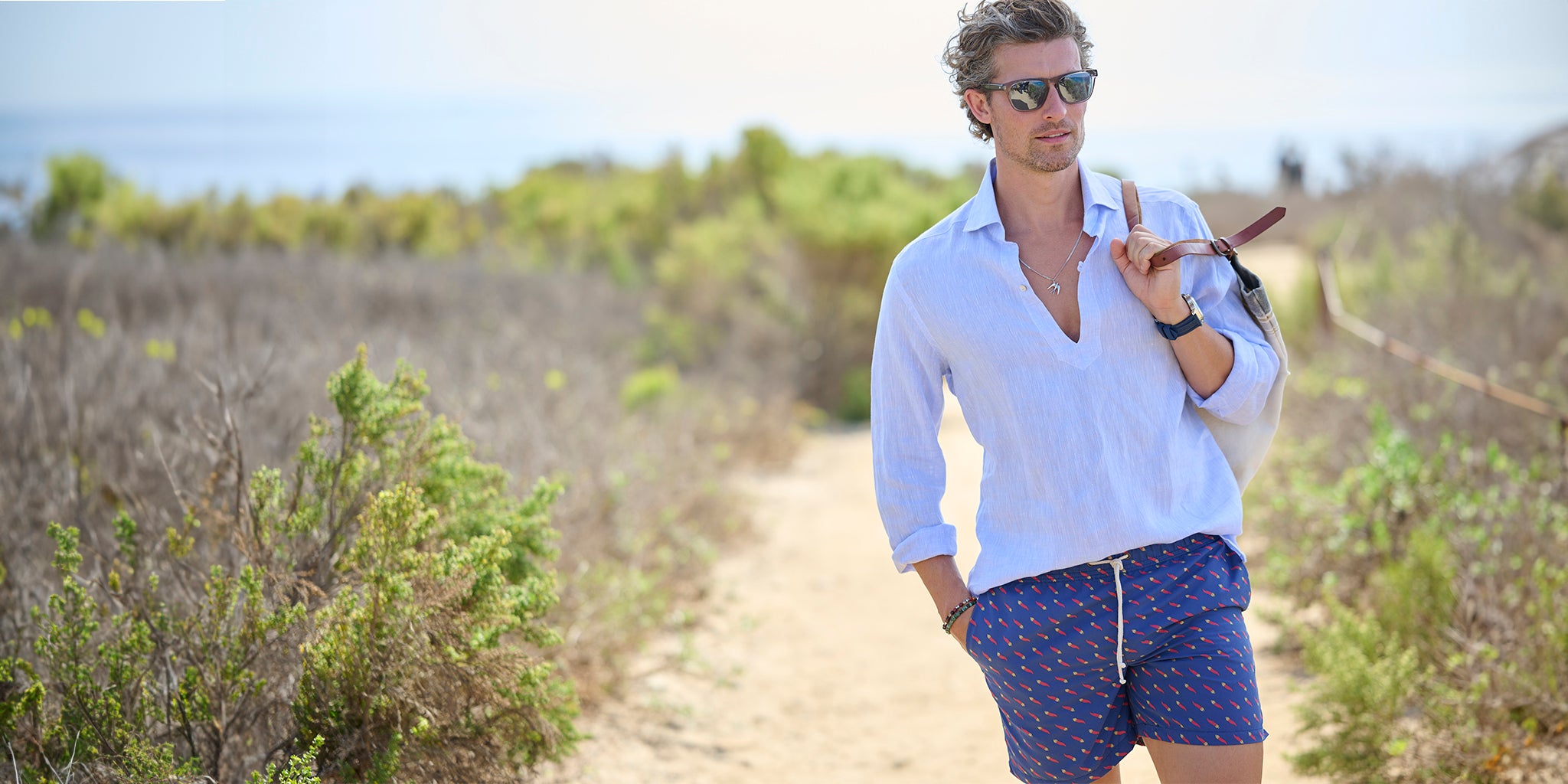 Man wearing a light blue shirt and patterned swim shorts, walking on a path with greenery and the ocean in the background.