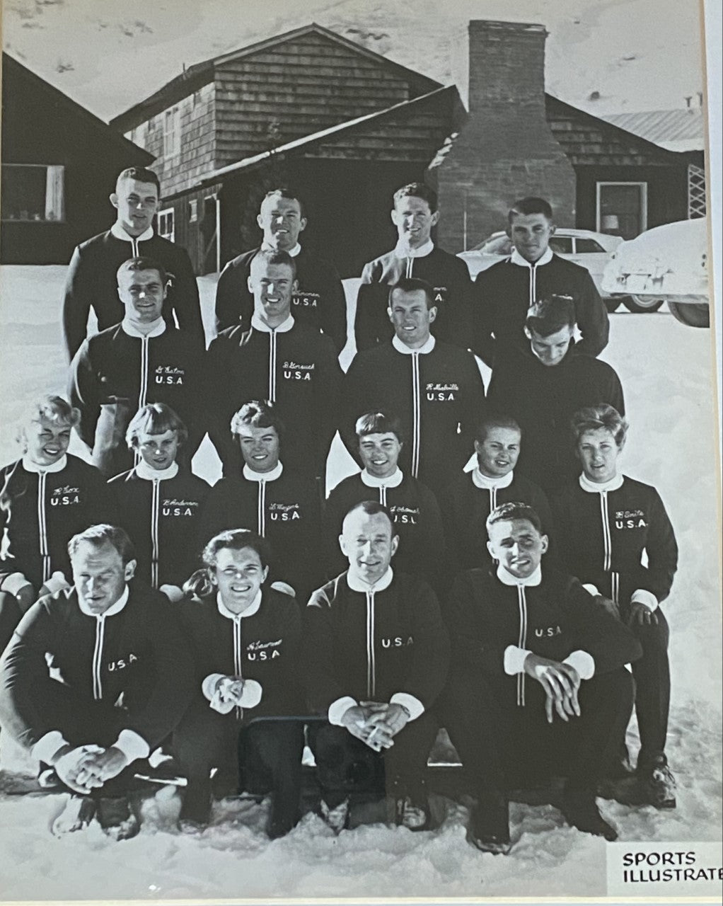 Men and Women from the US Ski Team in matching jackets posing outdoors with buildings and a car in the background.
